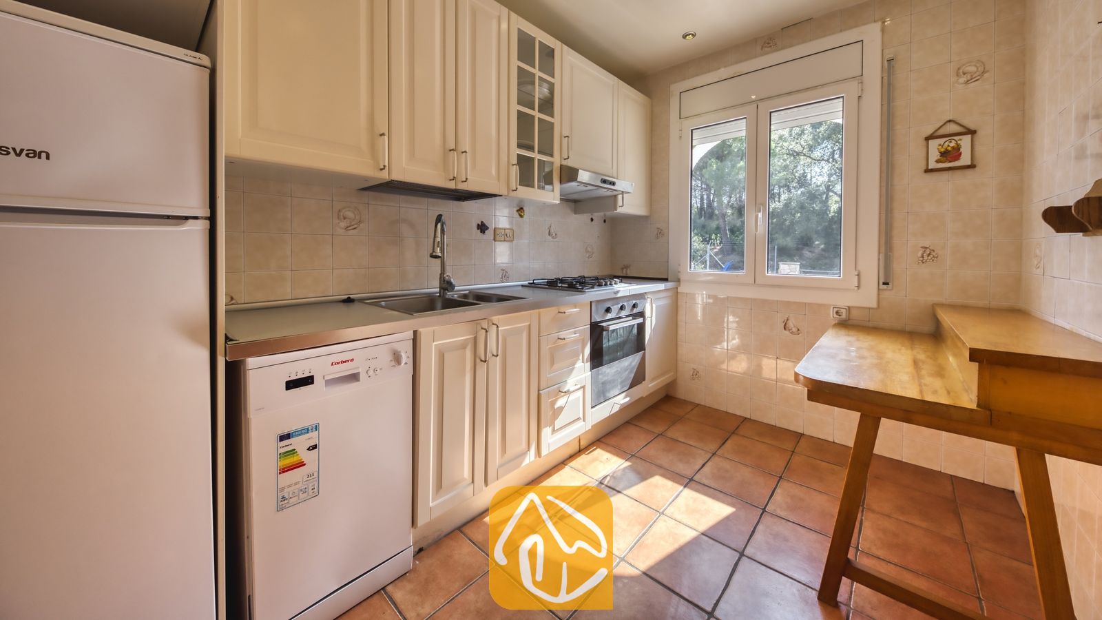 Vakantievilla's Costa Brava Spanje - Villa Nicky - Keuken met witte kasten, inbouwoven, vaatwasser, betegelde muren en een houten bartafel bij het raam.
