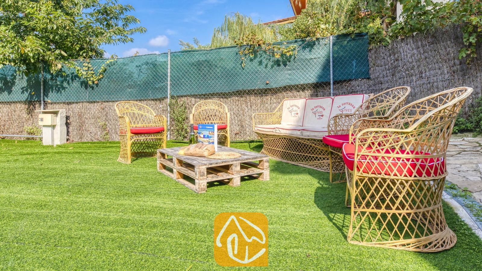 Vakantievilla's Costa Brava Spanje - Villa Nicky - Buitenlounge van Villa Nicky met rieten fauteuils, een bank met rood-witte kussens en een salontafel van houten pallets op kunstgras.