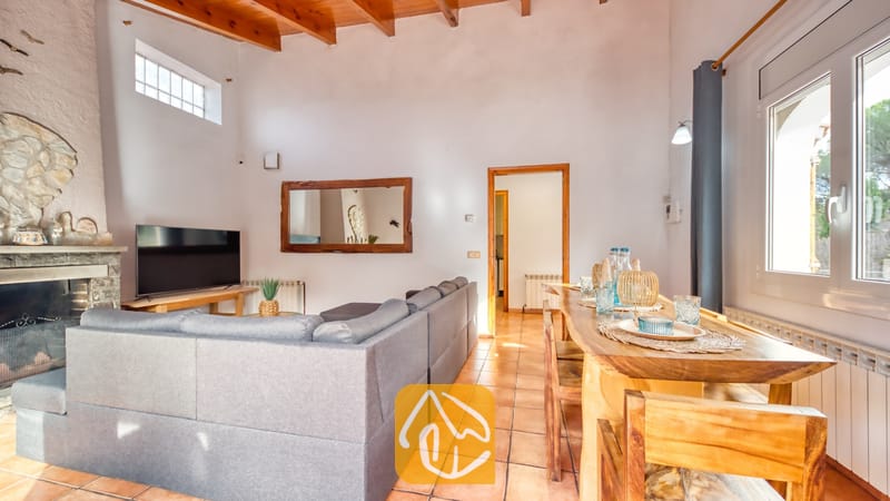 Vakantievilla's Costa Brava Spanje - Villa Nicky - Woon- en eetgedeelte met een grijze hoekbank, een houten eettafel, tegelvloer, authentieke houten balken aan het plafond en een open haard.