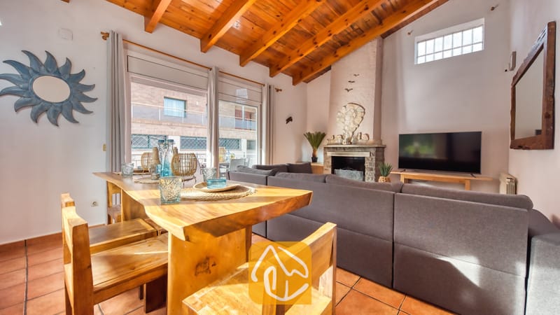 Vakantievilla's Costa Brava Spanje - Villa Nicky - Woonkamer binnen in Villa Nicky met een houten eettafel, een grote grijze bank, een open haard en authentieke houten plafondbalken.