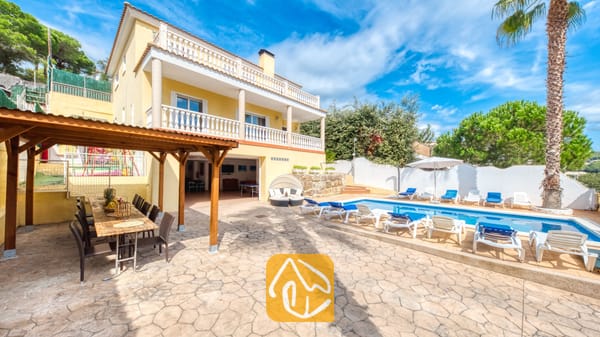 Vakantievilla's Costa Brava Spanje - Villa Ashley - Privézwembad bij Villa Ashley met helderblauw water en witte ligbedden. De gele villa is voorzien van balkons met witte balustrades en een houten pergola voor heerlijke diners in de buitenlucht op het stenen terras.