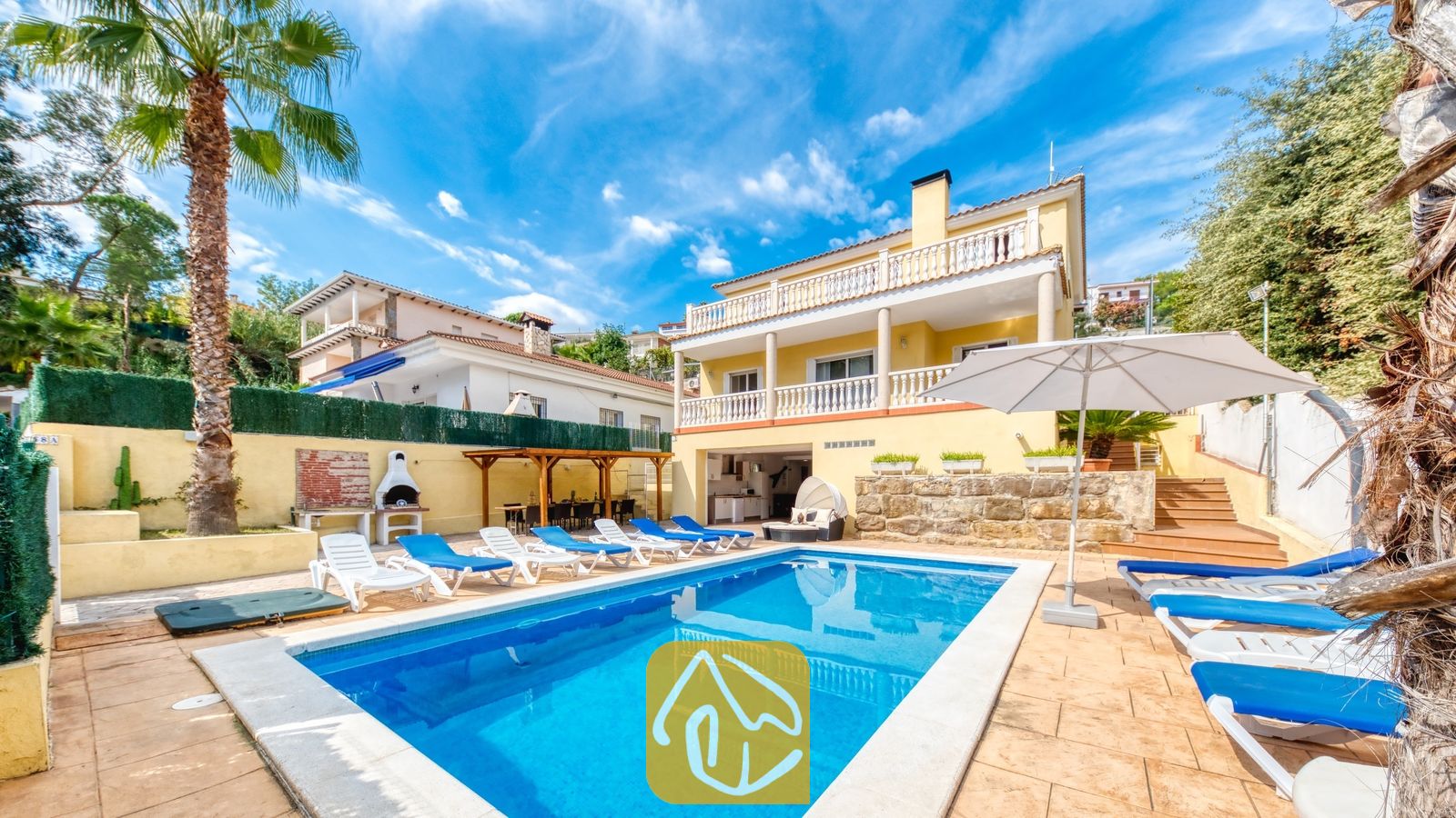 Vakantievilla's Costa Brava Spanje - Villa Ashley - Privézwembad bij Villa Ashley, omringd door blauwe ligbedden, een witte parasol en een geel gebouw van twee verdiepingen met witte balkonhekken.