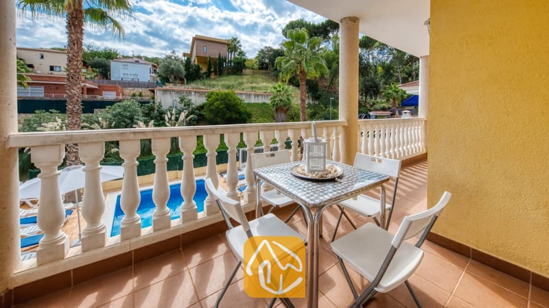 Vakantievilla's Costa Brava Spanje - Villa Ashley - Terras van Villa Ashley met een metalen tafel, witte stoelen en een stenen balustrade met uitzicht op een blauw zwembad en palmbomen.