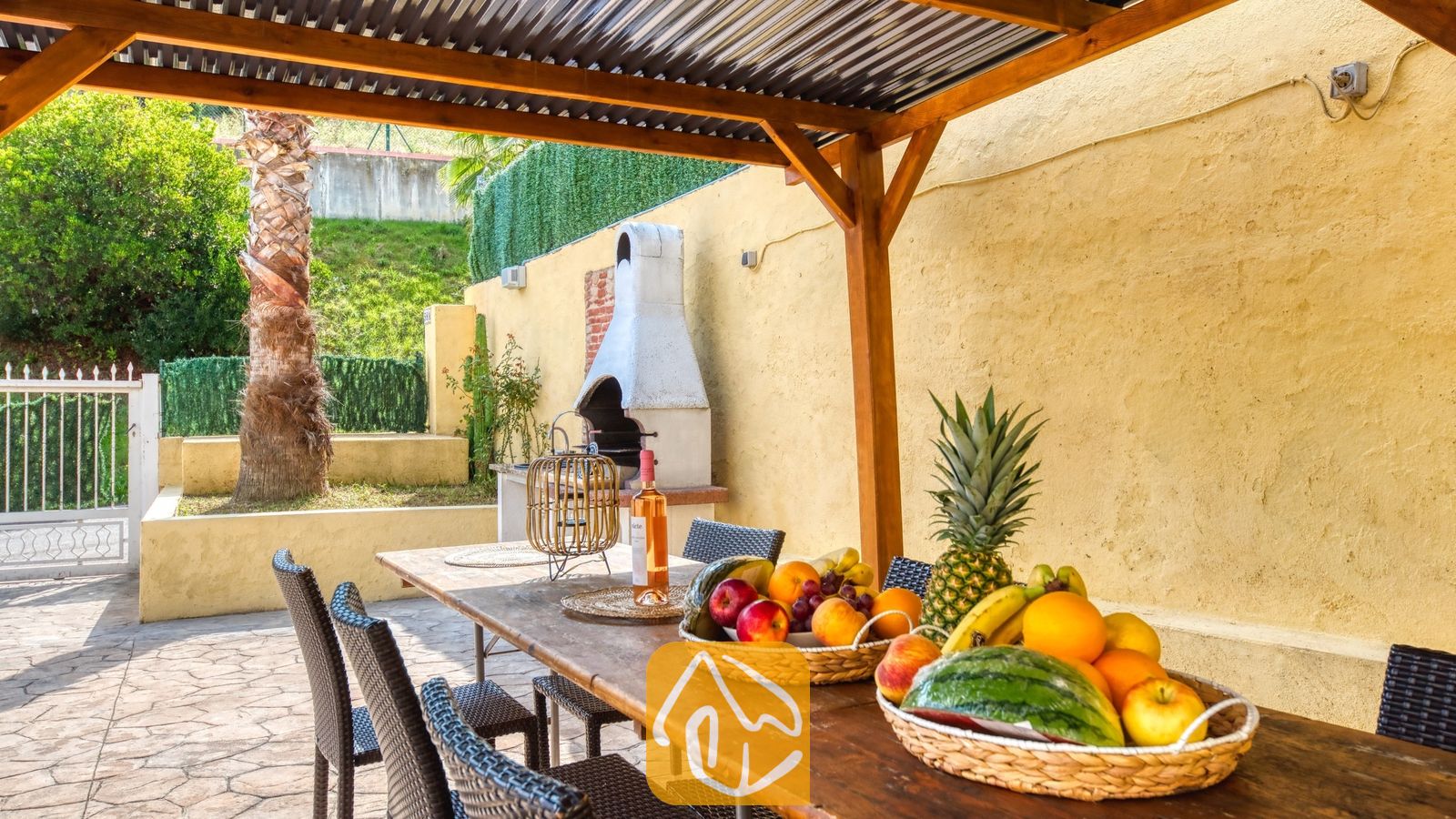 Vakantievilla's Costa Brava Spanje - Villa Ashley - Houten eettafel onder een pergola bij Villa Ashley, gedekt met vers fruit en wijn. Een witte buitenhaard staat tegen een gele muur nabij een palmboom.
