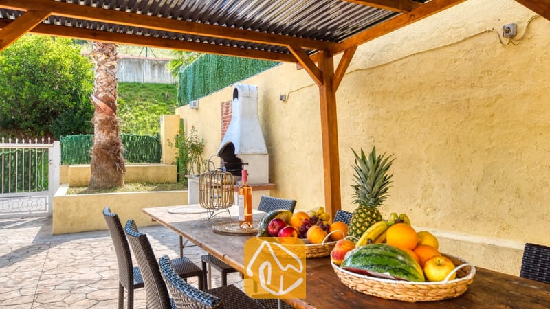 Vakantievilla's Costa Brava Spanje - Villa Ashley - Houten eettafel onder een pergola bij Villa Ashley, gedekt met vers fruit en wijn. Een witte buitenhaard staat tegen een gele muur nabij een palmboom.