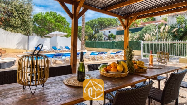 Vakantievilla's Costa Brava Spanje - Villa Ashley - Houten eettafel onder een pergola bij Villa Ashley, voorzien van fruitmanden en lantaarns, met uitzicht op het buitenzwembad en de ligbedden.