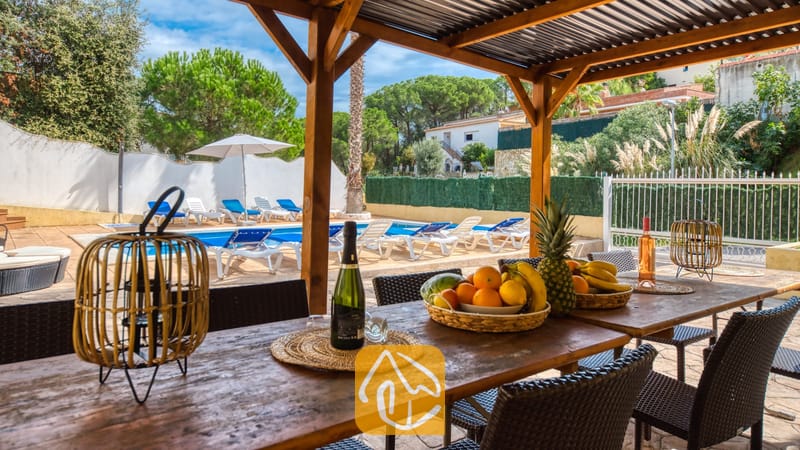 Vakantievilla's Costa Brava Spanje - Villa Ashley - Houten eettafel onder een pergola bij Villa Ashley, voorzien van fruitmanden en lantaarns, met uitzicht op het buitenzwembad en de ligbedden.