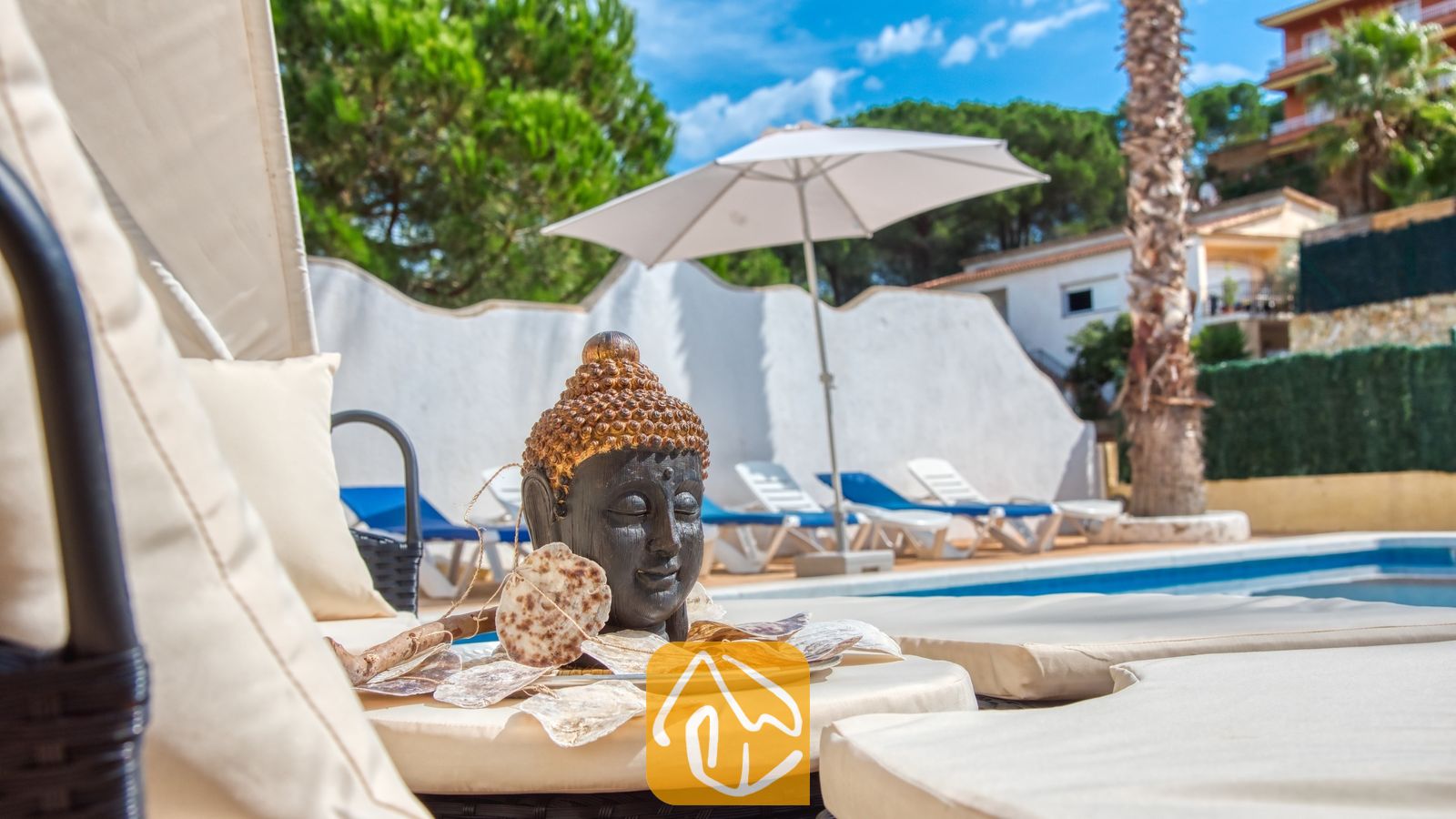 Vakantievilla's Costa Brava Spanje - Villa Ashley - Decoratief Boeddhabeeld op een crèmekleurig ligbed bij Villa Ashley, met op de achtergrond het zwembad, witte ligstoelen en een grote parasol.
