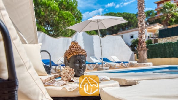 Vakantievilla's Costa Brava Spanje - Villa Ashley - Decoratief Boeddhabeeld op een crèmekleurig ligbed bij Villa Ashley, met op de achtergrond het zwembad, witte ligstoelen en een grote parasol.