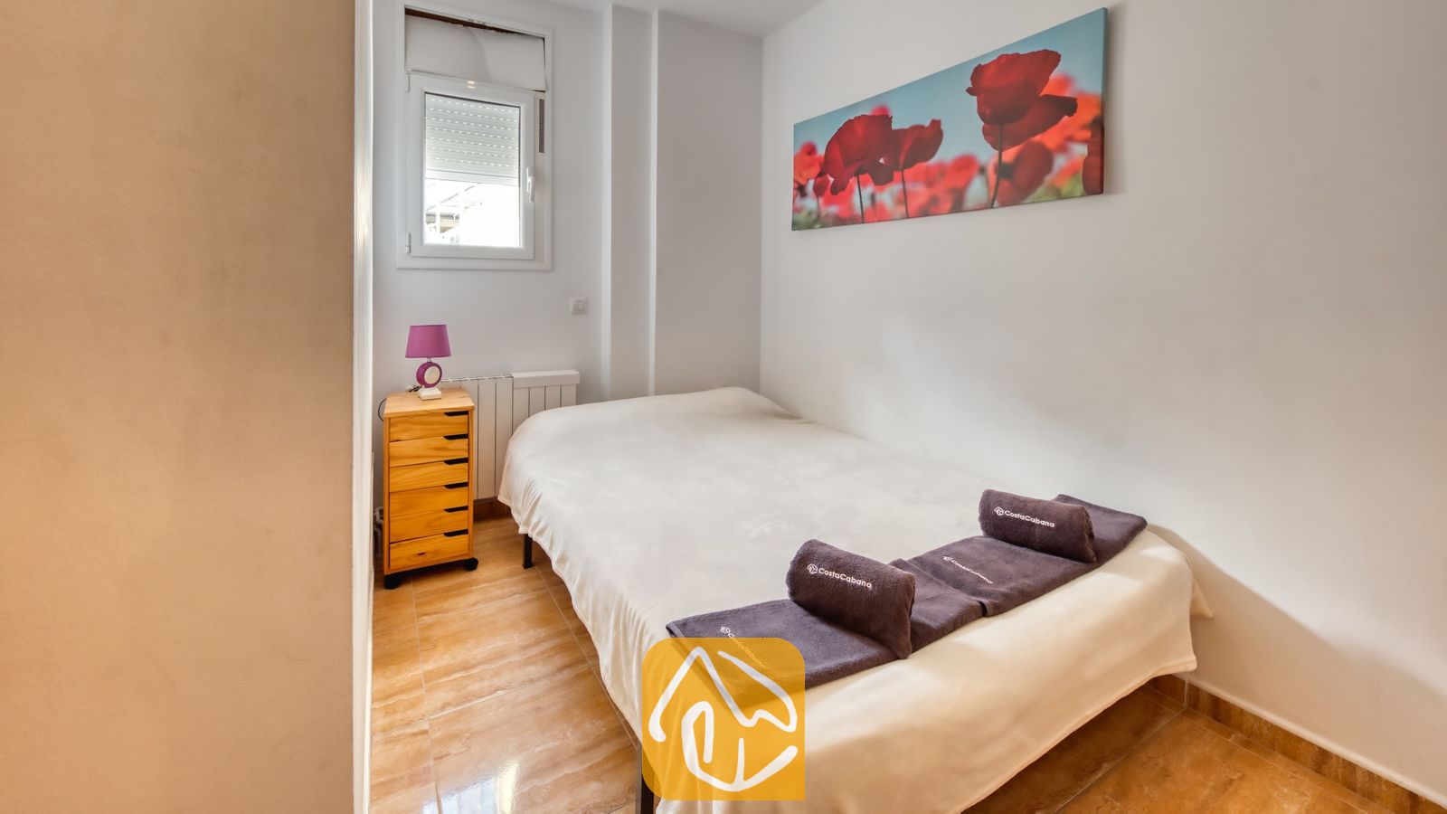 Vakantievilla's Costa Brava Spanje - Villa Ashley - Eenpersoonsbed met wit beddengoed en paarse handdoeken in een slaapkamer van Villa Ashley. Een houten ladekast met een paarse lamp staat bij het raam, naast kunstwerken met klaprozen.