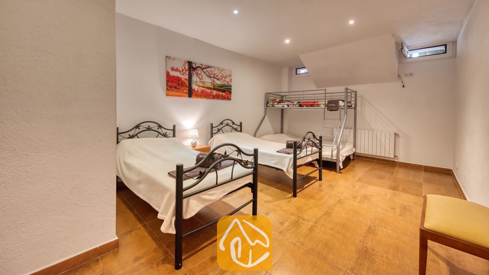 Vakantievilla's Costa Brava Spanje - Villa Ashley - Slaapkamer met twee eenpersoonsbedden van zwart metaal en een stapelbed in Villa Ashley. De kamer is voorzien van een warme tegelvloer, witte muren en een kleurrijk landschapsschilderij.
