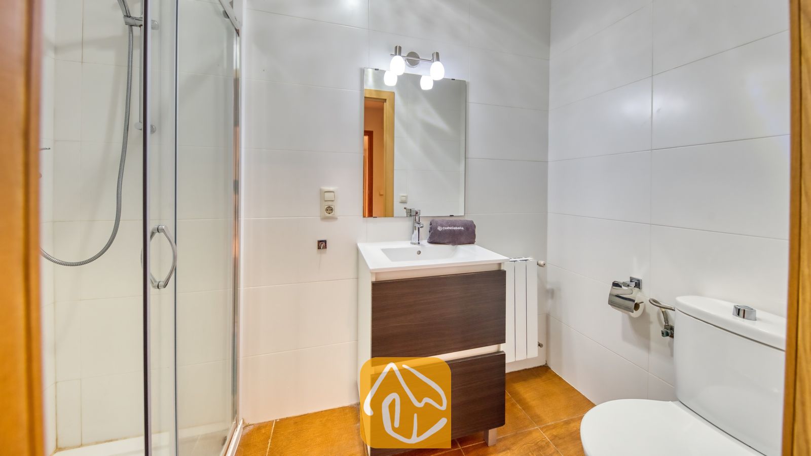 Vakantievilla's Costa Brava Spanje - Villa Ashley - Badkamer met een wastafelmeubel van donker hout, een inloopdouche met glazen wand en wit betegelde muren. Boven de spiegel is een verlichtingsarmatuur met drie lampen gemonteerd.