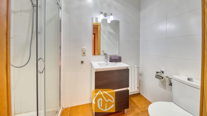 Villas de vacances Costa Brava Espagne - Villa Ashley - Salle de bain équipée d'un meuble-lavabo en bois sombre, d'une douche vitrée et de murs en carrelage blanc. Un luminaire à trois ampoules est installé au-dessus du miroir.