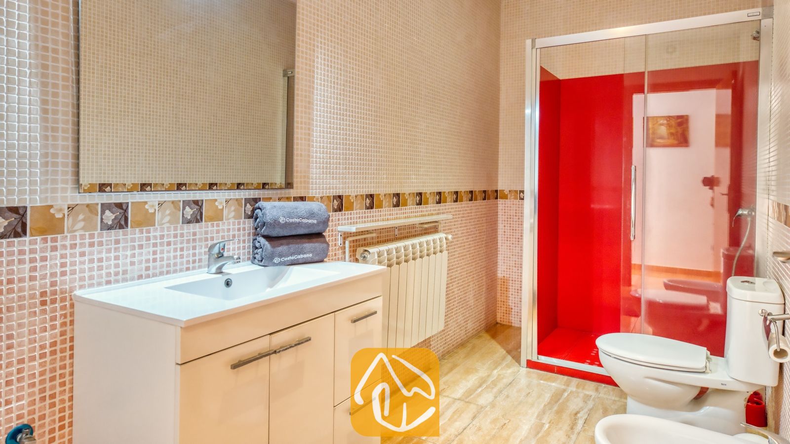 Vakantievilla's Costa Brava Spanje - Villa Ashley - Badkamermeubel met een witte wastafel en grijze handdoeken naast een inloopdouche met een felrode accentmuur en een glazen wand.