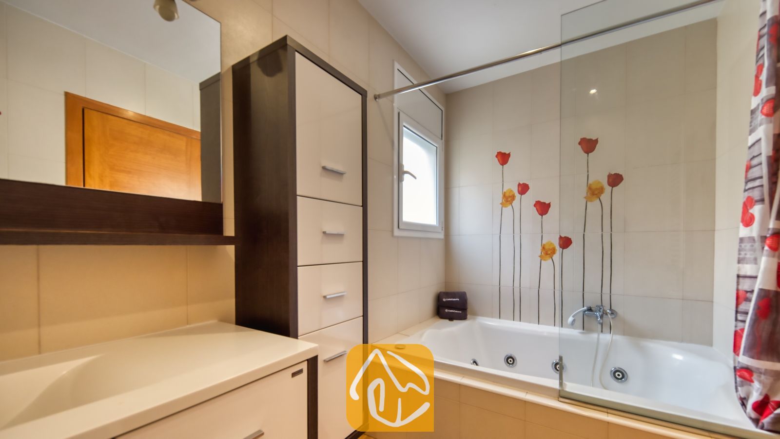 Vakantievilla's Costa Brava Spanje - Villa Ashley - Whirlpoolbad met glazen wand en bloemrijke muurdecoratie in rode tinten. De kamer is voorzien van een hoge opbergkast met witte lades en een kaptafelspiegel waarin een houten deur weerspiegelt.