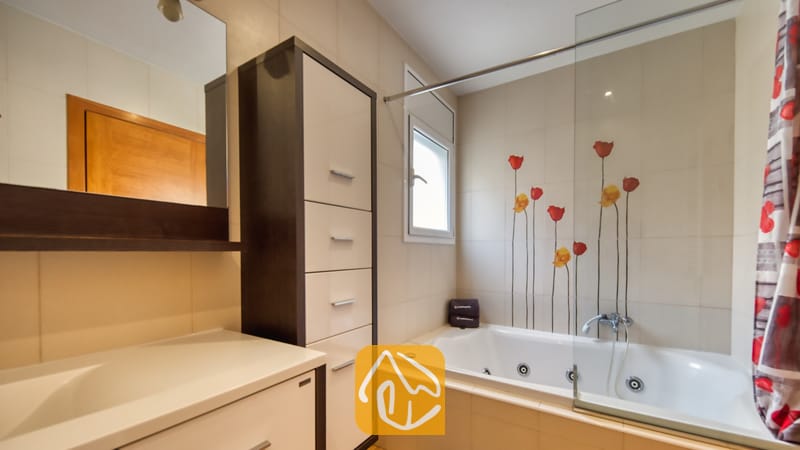 Villas de vacances Costa Brava Espagne - Villa Ashley - Baignoire balnéo avec paroi vitrée et décoration murale florale rouge. La pièce comprend une colonne de rangement haute dotée de tiroirs blancs, ainsi qu'un miroir de courtoisie reflétant une porte en bois.