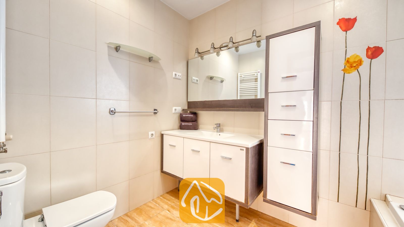 Vakantievilla's Costa Brava Spanje - Villa Ashley - Badkamermeubel met witte kasten, een grote spiegel met bovenverlichting en een hoge opbergkast naast een muur met rode en gele bloemmotieven.