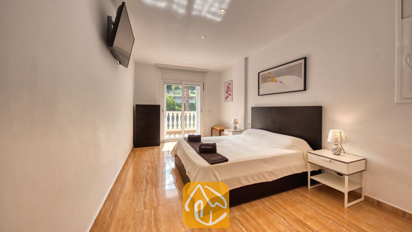 Vakantievilla's Costa Brava Spanje - Villa Ashley - Slaapkamer in Villa Ashley met een tweepersoonsbed, een donker hoofdbord en wit beddengoed. De kamer is voorzien van een flatscreen-tv aan de muur, glanzende tegelvloeren en een deur naar het balkon.