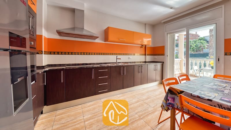 Vakantievilla's Costa Brava Spanje - Villa Ashley - De keuken van Villa Ashley is voorzien van donkerhouten onderkasten, stijlvolle oranje accenten aan de bovenzijde en een eettafel met bijpassende oranje stoelen bij de glazen terrasdeur.