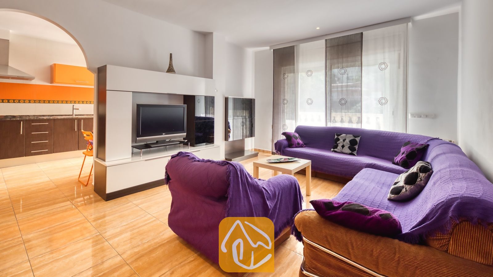 Vakantievilla's Costa Brava Spanje - Villa Ashley - Paarse banken in de woonkamer van Villa Ashley, gecombineerd met een wit tv-meubel, glanzende tegelvloeren en een boogdoorgang die leidt naar een keuken met oranje en bruine kasten.