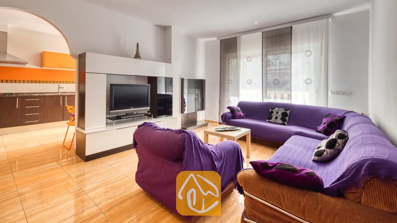 Vakantievilla's Costa Brava Spanje - Villa Ashley - Paarse banken in de woonkamer van Villa Ashley, gecombineerd met een wit tv-meubel, glanzende tegelvloeren en een boogdoorgang die leidt naar een keuken met oranje en bruine kasten.