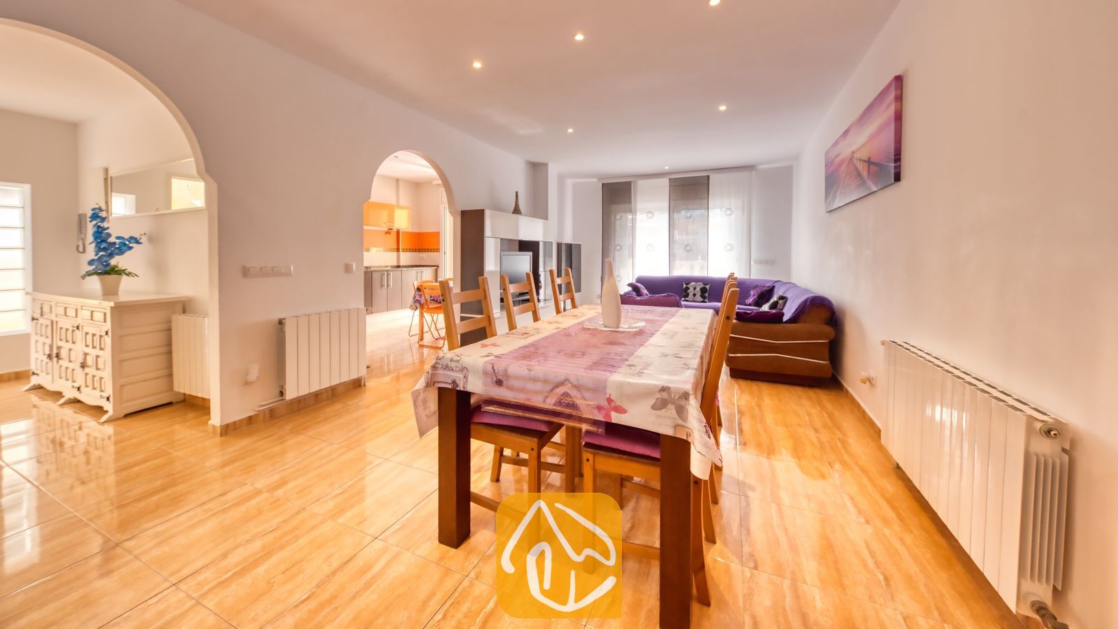 Vakantievilla's Costa Brava Spanje - Villa Ashley - De eethoek van Villa Ashley is voorzien van een houten tafel met een paars tafelkleed met patroon en bijpassende stoelen. De glanzende tegelvloer loopt naadloos over in de woonkamer, waar een comfortabele paarse bank staat.