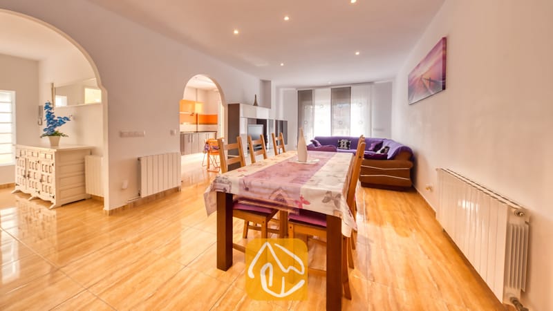 Vakantievilla's Costa Brava Spanje - Villa Ashley - De eethoek van Villa Ashley is voorzien van een houten tafel met een paars tafelkleed met patroon en bijpassende stoelen. De glanzende tegelvloer loopt naadloos over in de woonkamer, waar een comfortabele paarse bank staat.