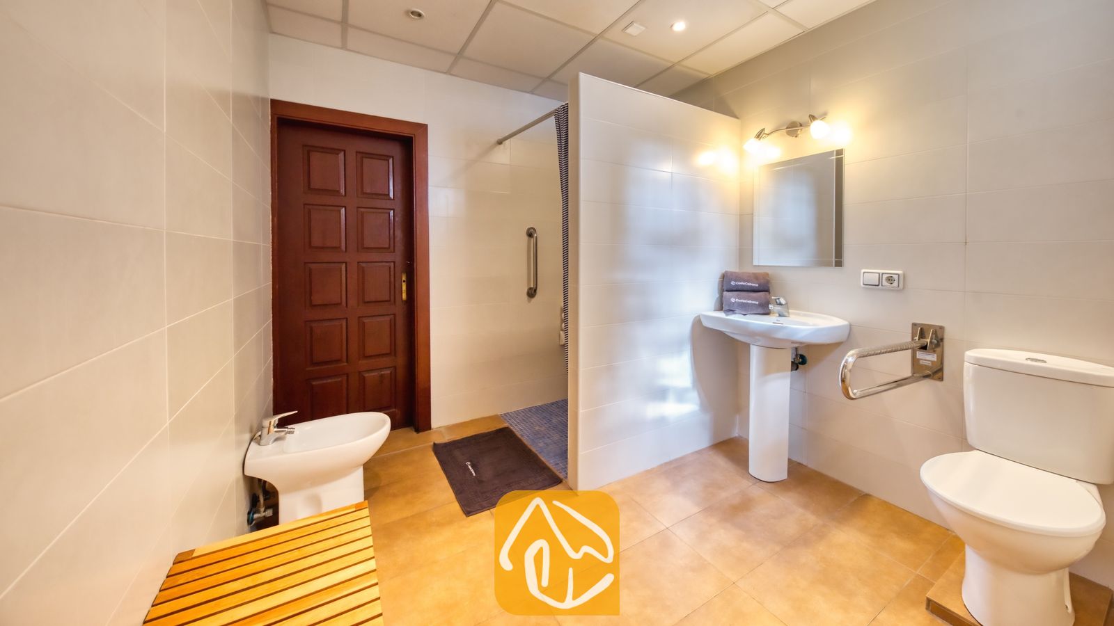 Vakantievilla's Costa Brava Spanje - Villa Ashley - Toegankelijke badkamer met inloopdouche voorzien van een wandbeugel, een witte wastafel op zuil en een toilet met steunbeugel. De ruimte is afgewerkt met beige vloertegels en witte muren.