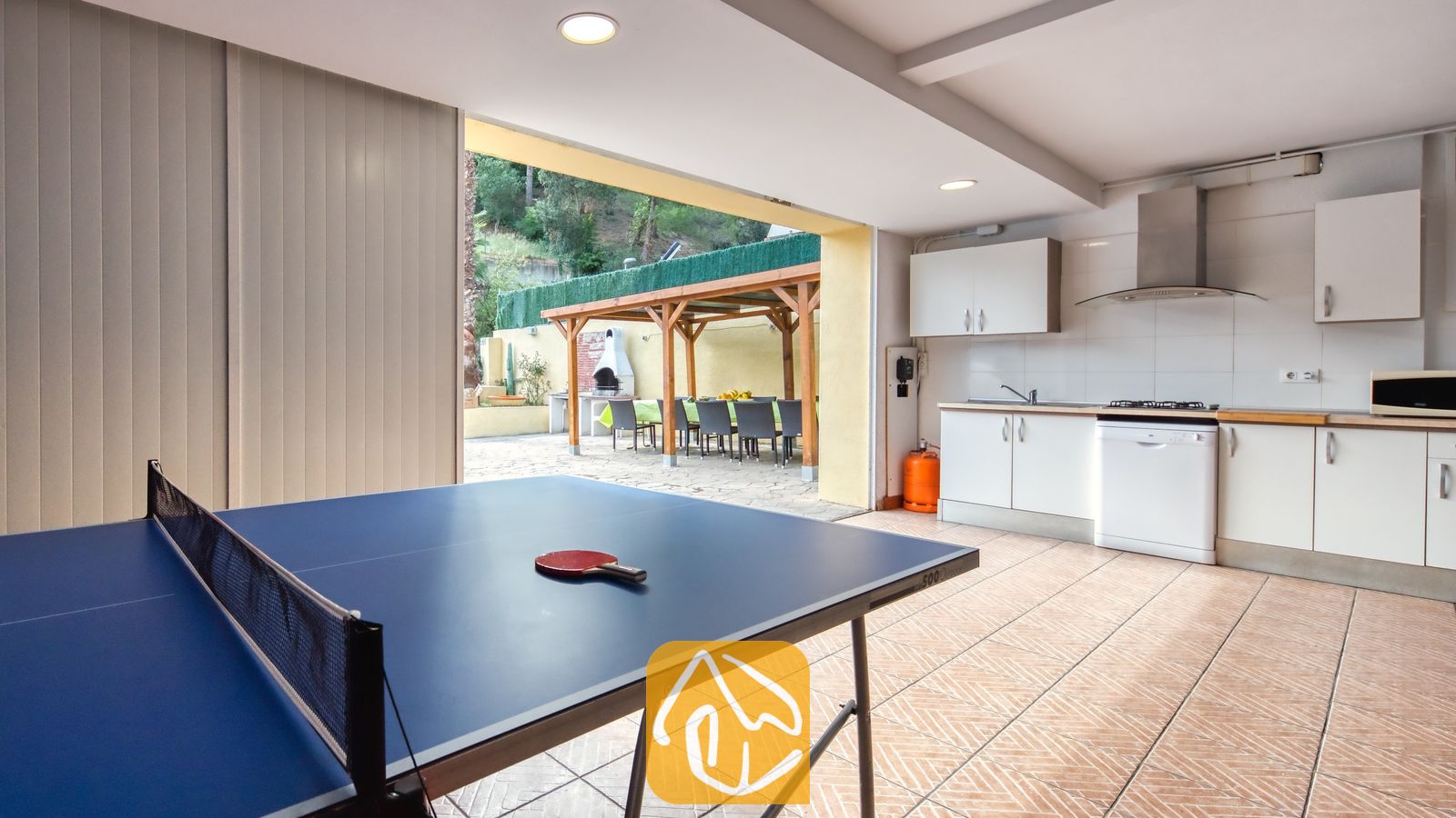 Vakantievilla's Costa Brava Spanje - Villa Ashley - Tafeltennistafel met een rood batje in een betegelde ruimte van Villa Ashley. De ruimte beschikt over een witte keuken en biedt directe toegang tot een terras met een houten pergola en een stenen barbecue.