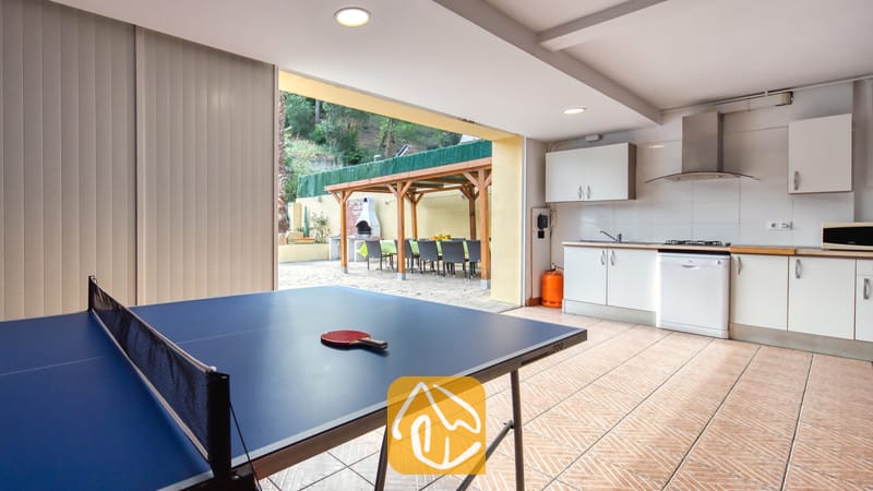 Villas de vacances Costa Brava Espagne - Villa Ashley - Table de ping-pong avec raquette rouge dans une pièce carrelée à la Villa Ashley. L'espace comprend une cuisine blanche et s'ouvre sur une terrasse agrémentée d'une pergola en bois et d'un barbecue en pierre.