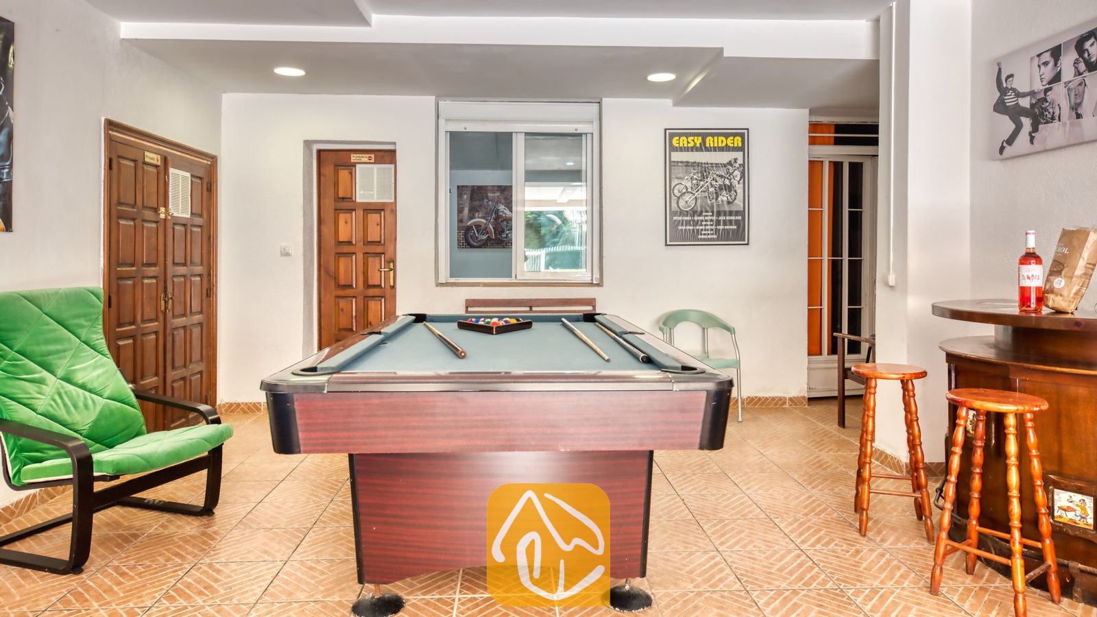 Vakantievilla's Costa Brava Spanje - Villa Ashley - Biljarttafel met blauw laken in de recreatieruimte van Villa Ashley, voorzien van een houten bar met barkrukken, een groene fauteuil en een tegelvloer.