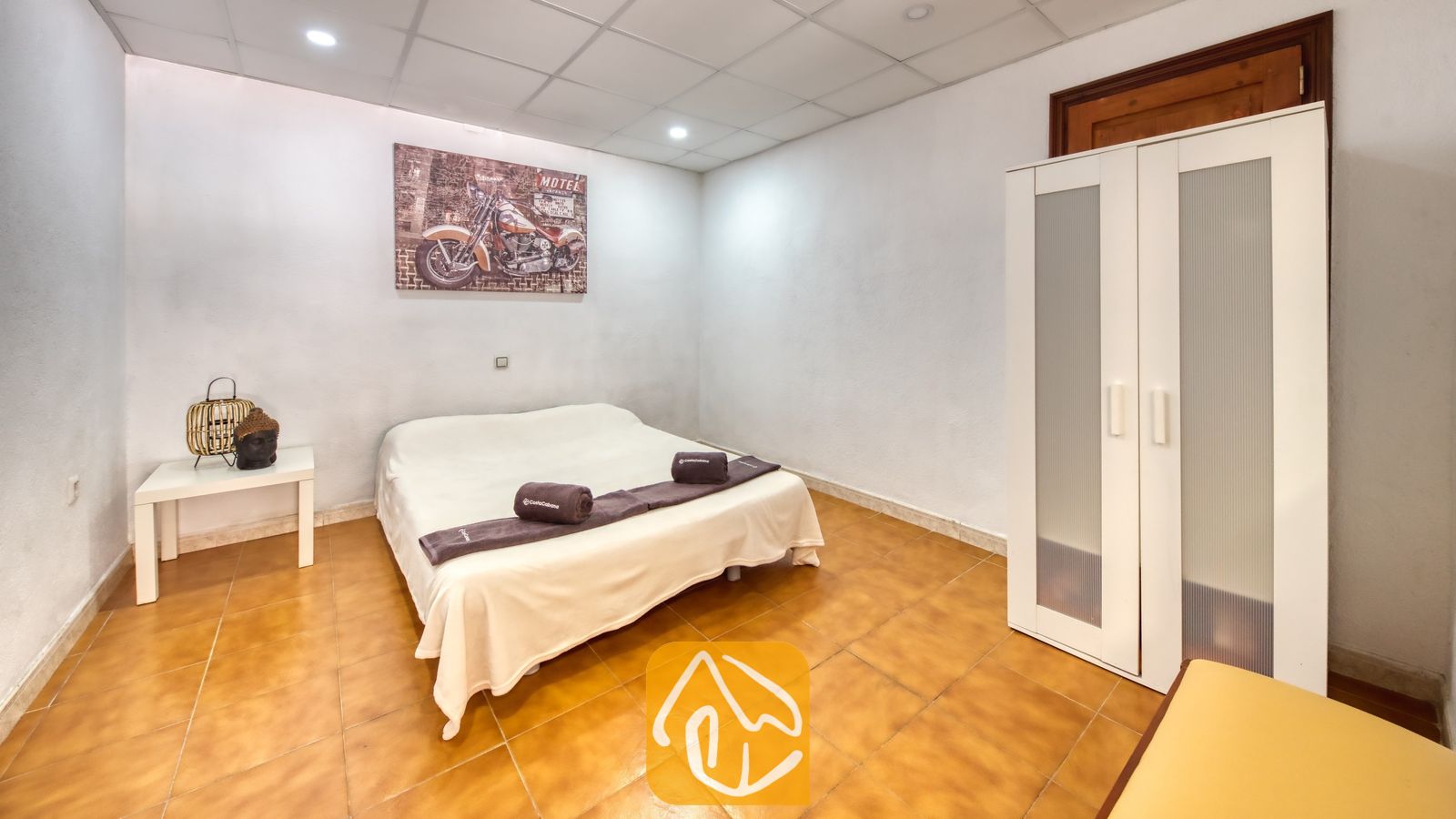 Vakantievilla's Costa Brava Spanje - Villa Ashley - Slaapkamer in Villa Ashley met een tweepersoonsbed, wit beddengoed en paarse handdoeken. De kamer is voorzien van terracotta plavuizen, een witte kledingkast en muurdecoratie met een motorthema.