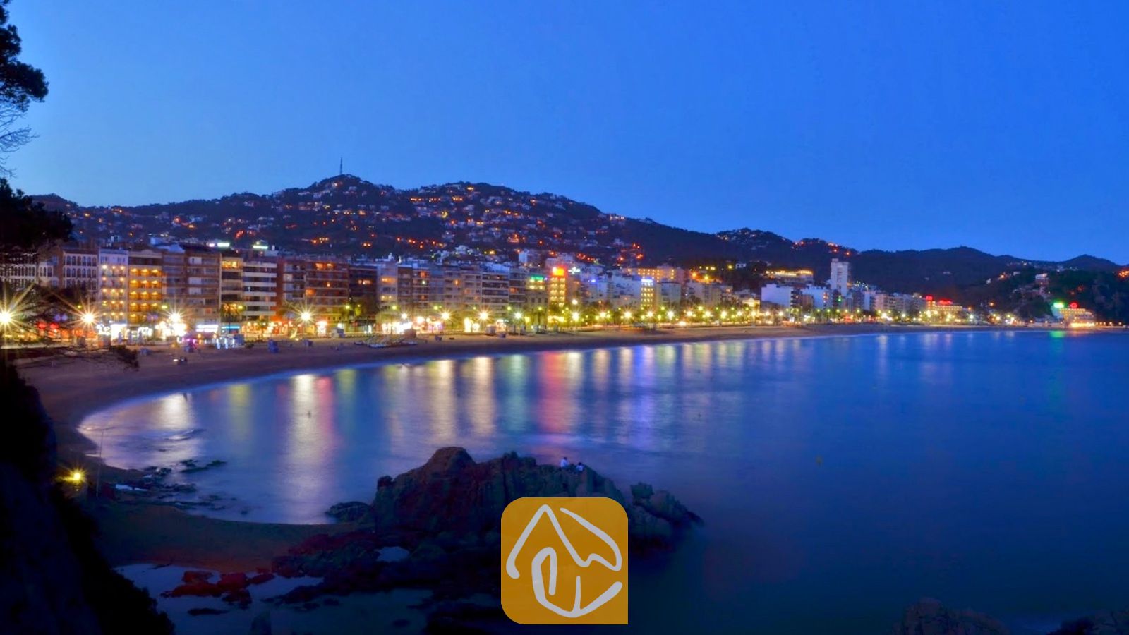 Vakantievilla's Costa Brava Spanje - Villa Pilar - Kustplaats en zandstrand verlicht in de schemering met kleurrijke lichtjes die weerspiegelen op het kalme water, gezien vanuit Villa Pilar.