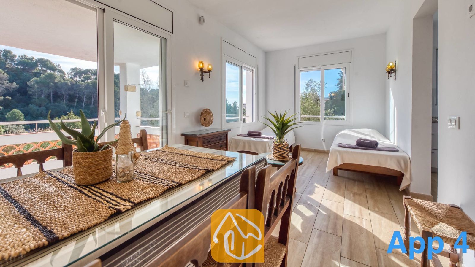 Vakantievilla's Costa Brava Spanje - Villa Pilar - Lichte eet- en slaapkamer in Villa Pilar, voorzien van een houten tafel met glazen blad en geweven placemat, tegelvloeren en grote ramen met uitzicht naar buiten.