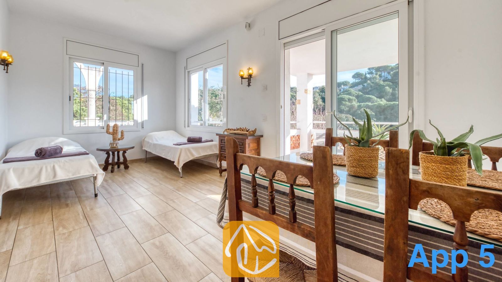 Vakantievilla's Costa Brava Spanje - Villa Pilar - Lichte multifunctionele ruimte in Villa Pilar met een houten eettafel op de voorgrond, een tegelvloer, twee eenpersoonsbedden met wit beddengoed en grote ramen met uitzicht op het groen.