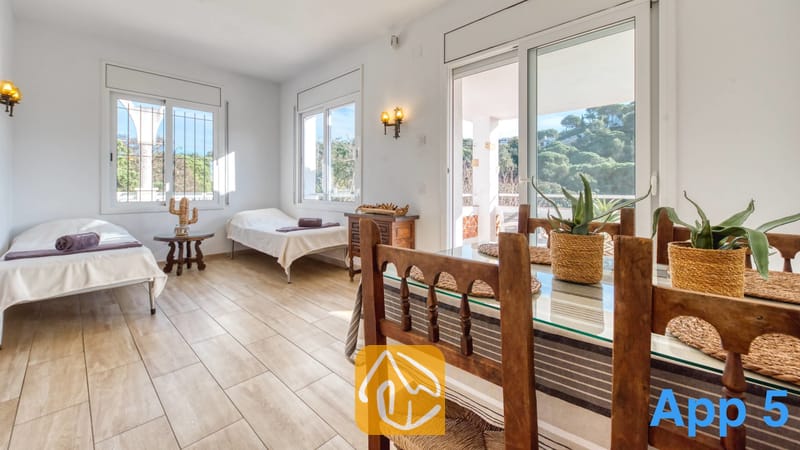 Villas de vacances Costa Brava Espagne - Villa Pilar - Lumineuse pièce polyvalente à la Villa Pilar, comprenant une table à manger en bois au premier plan, un sol carrelé, deux lits jumeaux avec du linge de lit blanc, et de grandes fenêtres offrant une vue sur la verdure.
