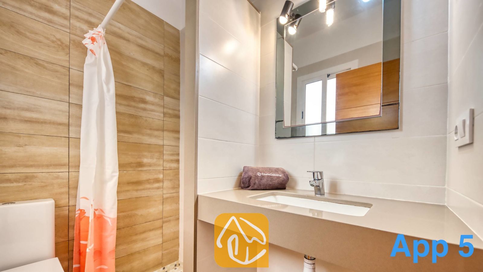 Vakantievilla's Costa Brava Spanje - Villa Pilar - Moderne badkamer met een wastafelmeubel van beige natuursteen, een vierkante wandspiegel met bovenverlichting en een inloopdouche met een wit-met-oranje geprint douchegordijn.
