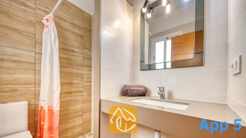 Villas de vacances Costa Brava Espagne - Villa Pilar - Salle de bains moderne dotée d'un meuble sous-vasque en pierre beige, d'un miroir mural carré avec éclairage intégré et d'une douche à l'italienne ornée d'un rideau à motifs blancs et orange.