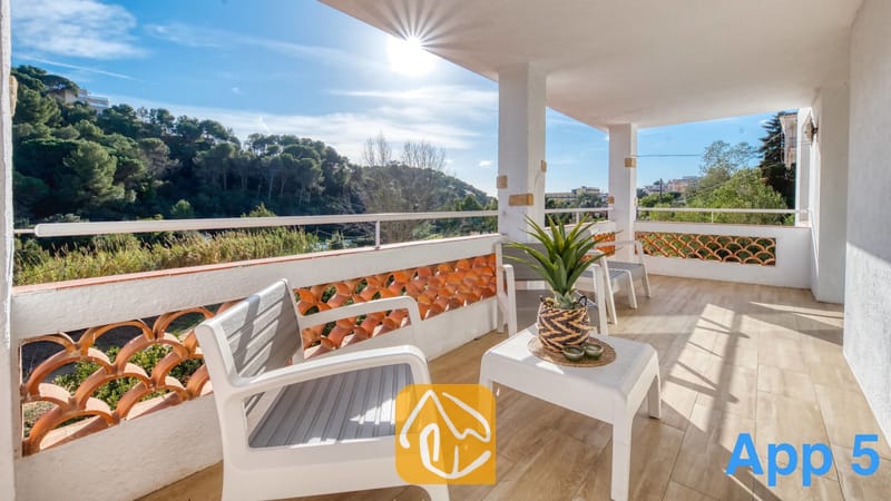 Villas de vacances Costa Brava Espagne - Villa Pilar - Terrasse couverte de la Villa Pilar avec des chaises d'extérieur blanches, une petite table supportant une plante en pot et un garde-corps doté de découpes en tuiles de terre cuite surplombant des collines verdoyantes.