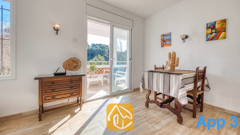 Villas de vacances Costa Brava Espagne - Villa Pilar - Lumineuse salle à manger de la Villa Pilar, dotée d'une table en bois, d'une nappe à rayures et d'une porte-fenêtre coulissante s'ouvrant sur une terrasse avec vue sur la forêt.