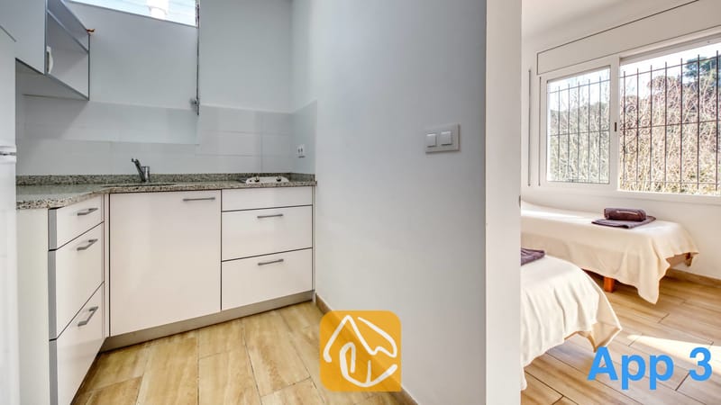 Villas de vacances Costa Brava Espagne - Villa Pilar - Coin cuisine compact équipé de meubles blancs, de plans de travail en granit et d'un évier en acier inoxydable, ouvert sur une chambre dotée de deux lits simples.