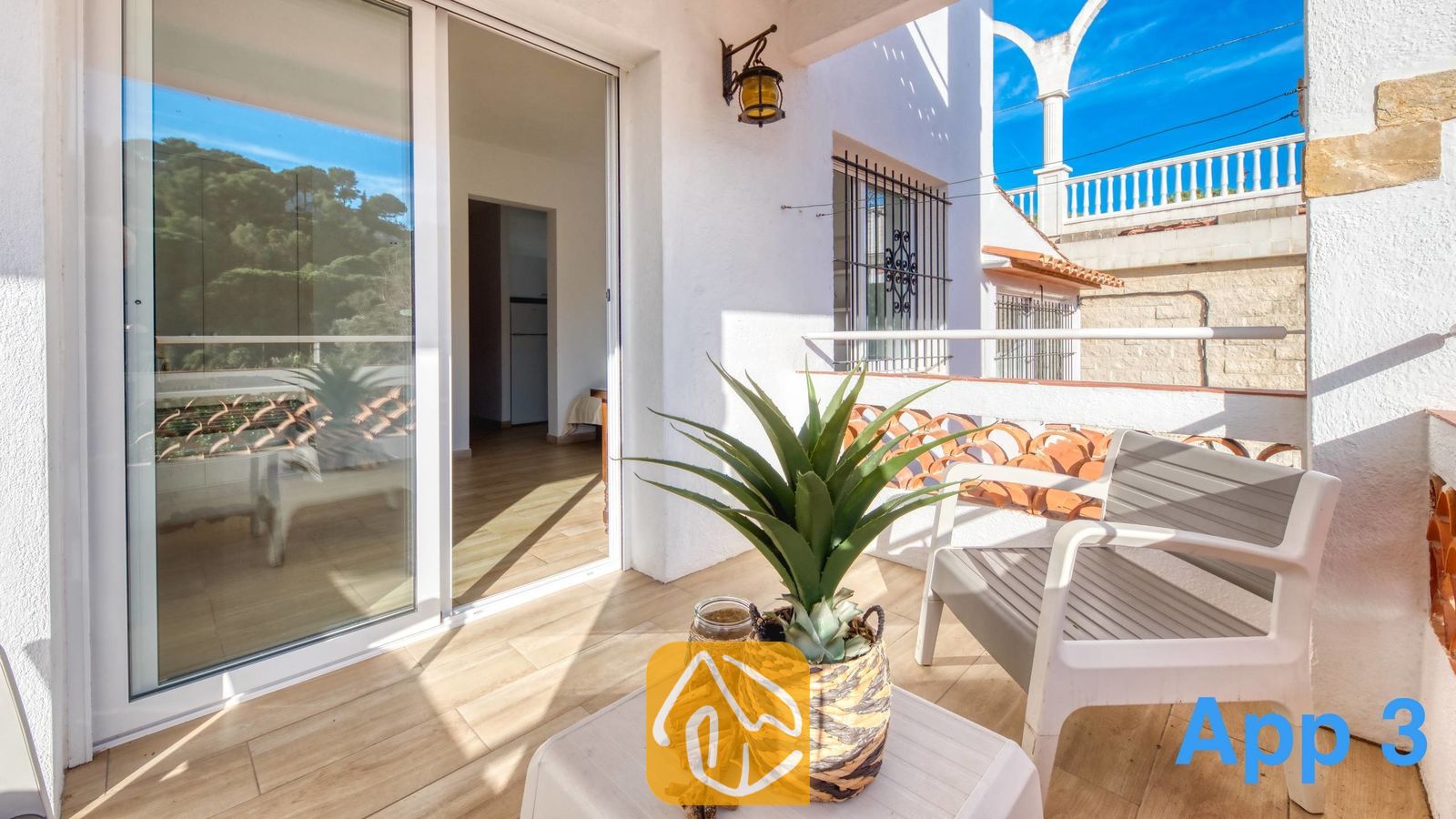 Vakantievilla's Costa Brava Spanje - Villa Pilar - Zonnig balkon bij Villa Pilar, voorzien van een witte fauteuil, een tafeltje met een plant in pot en een glazen schuifpui die toegang biedt tot het interieur.