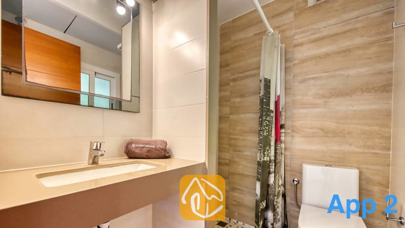 Villas de vacances Costa Brava Espagne - Villa Pilar - Salle de bain moderne dotée d'un plan de toilette beige avec vasque, d'un miroir rectangulaire mural et de toilettes situées à côté d'une douche pourvue d'un rideau à motifs.