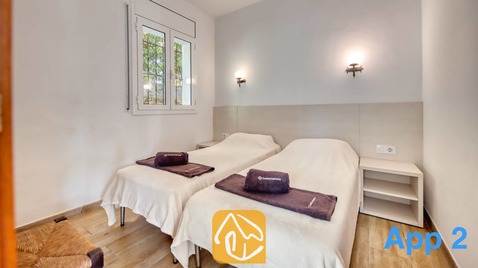 Vakantievilla's Costa Brava Spanje - Villa Pilar - Slaapkamer met twee eenpersoonsbedden voorzien van witte overtreksets, donker opgevouwen handdoeken, nachtkastjes en wandlampen onder een raam met dievenijzers.