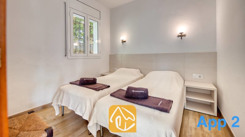 Villas de vacances Costa Brava Espagne - Villa Pilar - Chambre à lits jumeaux avec deux lits simples habillés de couvre-lits blancs, des serviettes sombres pliées, des tables de chevet et des appliques murales sous une fenêtre à barreau.