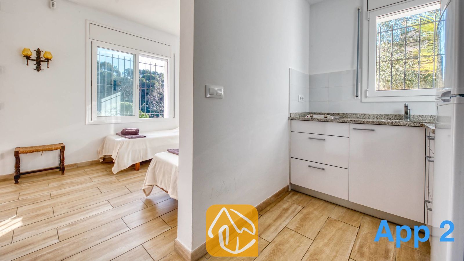 Vakantievilla's Costa Brava Spanje - Villa Pilar - Interieur van Villa Pilar met een compacte keuken voorzien van witte kasten, grenzend aan een slaapkamer met twee eenpersoonsbedden en houtlook tegelvloer.
