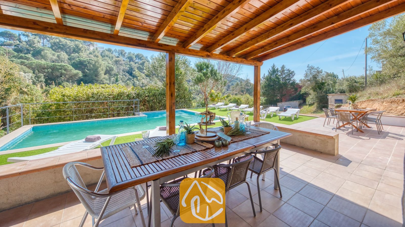 Vakantievilla's Costa Brava Spanje - Villa Pilar - Overdekte houten pergola bij Villa Pilar, voorzien van een eettafel buiten, een stenen terras en uitzicht op een zwembad en beboste heuvels.