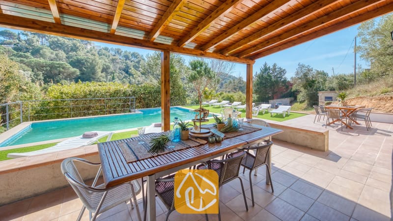 Vakantievilla's Costa Brava Spanje - Villa Pilar - Overdekte houten pergola bij Villa Pilar, voorzien van een eettafel buiten, een stenen terras en uitzicht op een zwembad en beboste heuvels.