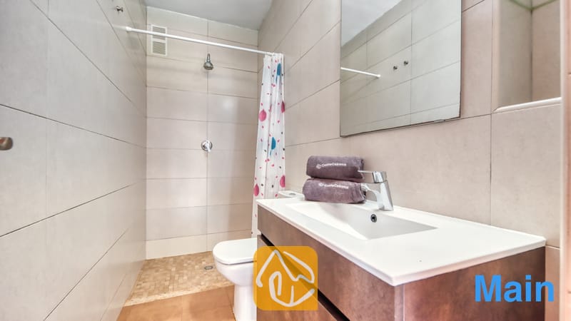 Villas de vacances Costa Brava Espagne - Villa Pilar - Salle de bains moderne dotée de murs carrelés de beige, d'un meuble sous-lavabo blanc avec des serviettes pliées, de toilettes et d'une douche à l'italienne pourvue d'un rideau à motifs.