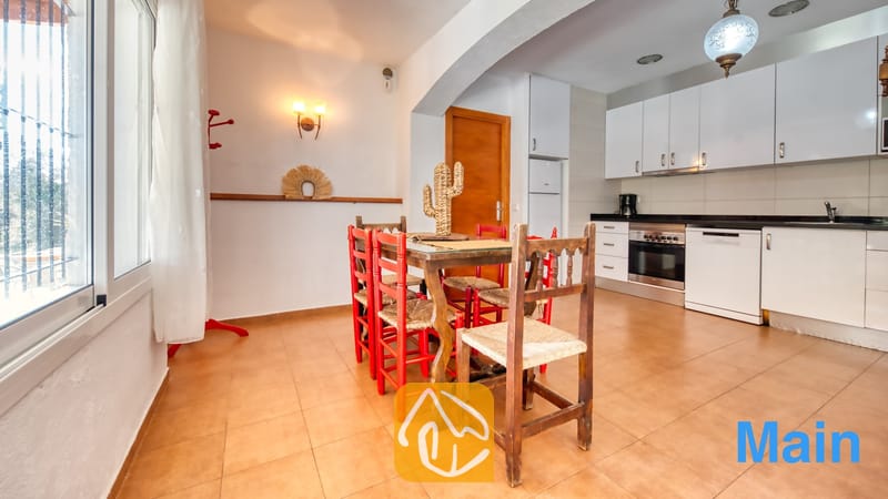 Vakantievilla's Costa Brava Spanje - Villa Pilar - Houten eettafel met rode en naturel biezen stoelen, geplaatst tussen een raam en een witte keuken bij Villa Pilar.